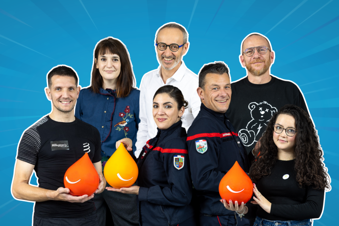 Groupe de sept adultes souriants posant ensemble sur un fond bleu. Certains portent des vêtements professionnels ou des uniformes, et plusieurs tiennent des objets en forme de goutte orange ou jaune.