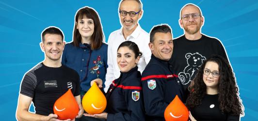 Groupe de sept adultes souriants posant ensemble sur un fond bleu. Certains portent des vêtements professionnels ou des uniformes, et plusieurs tiennent des objets en forme de goutte orange ou jaune.