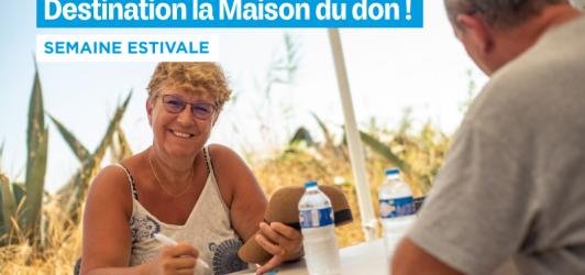 Une femme souriante aux cheveux blonds et portant des lunettes, assise à une table en plein air, elle rempli le questionnaire pré-don. Elle est en conversation avec un homme dont on ne voit que le dos. Le fond montre un champ de blé et un ciel bleu. En haut de l'image, un bandeau bleu affiche le texte « Destination la Maison du don ! » et en dessous, « SEMAINE ESTIVALE ».